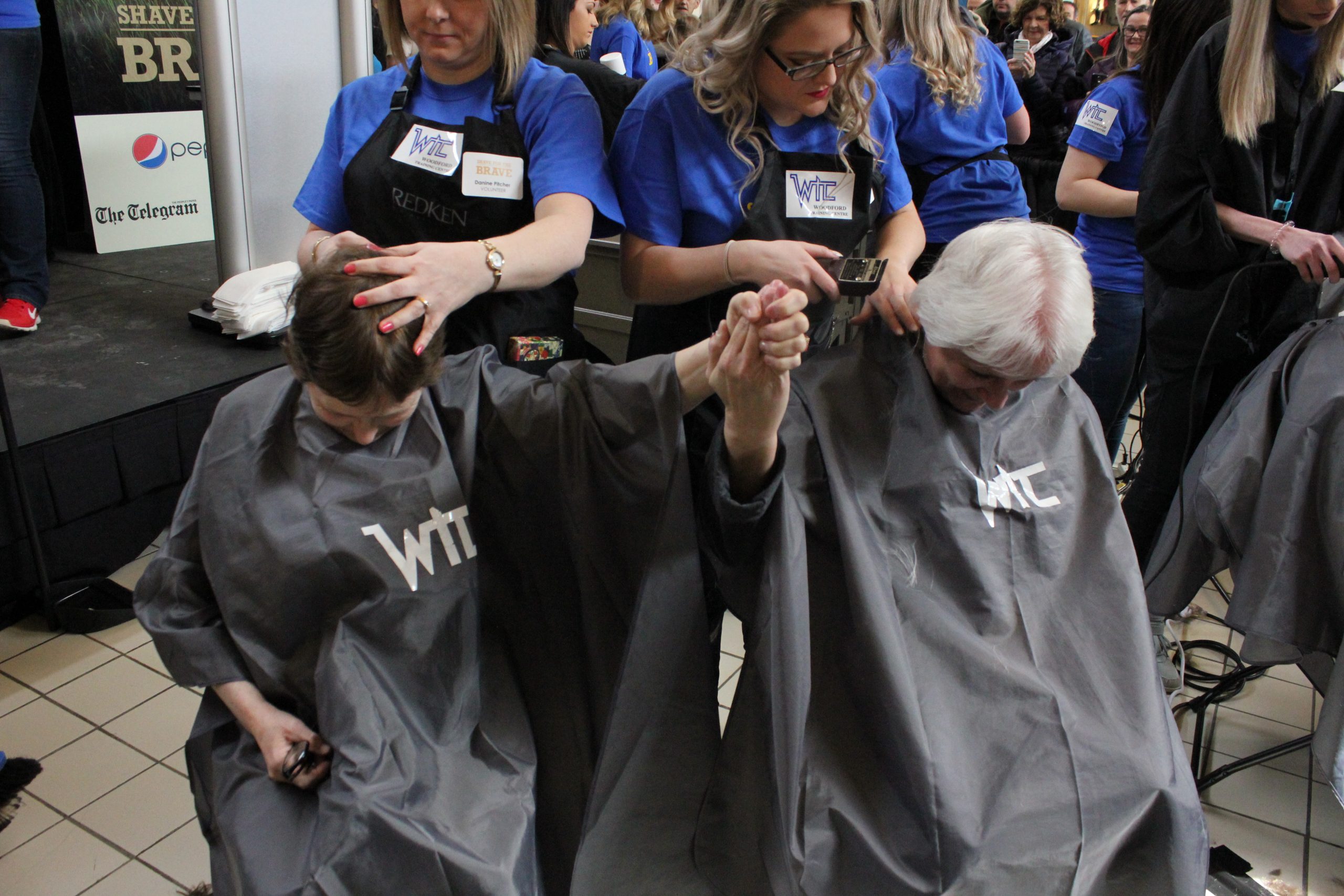 2 woman holding hands as they are about to get their heads shaved.