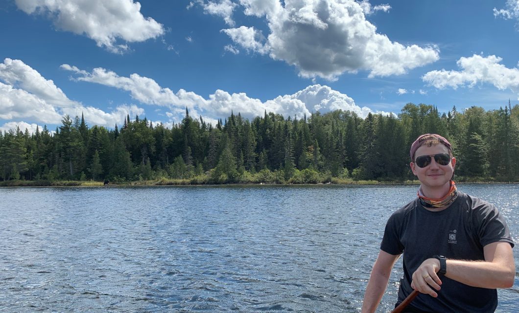 JR enjoying one of his favourite hobbies, canoe tripping in Algonquin Park. The sky is blue and filled with fluffy clouds. There is a line of forest in the middle with water below. JR is positioned at the right of the frame, wearing a dark t-shirt and sunglasses.
