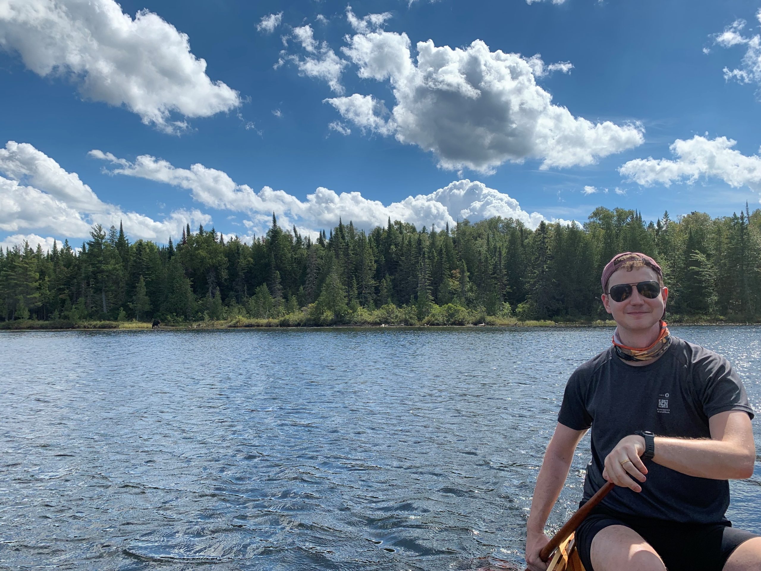 JR enjoying one of his favourite hobbies, canoe tripping in Algonquin Park. The sky is blue and filled with fluffy clouds. There is a line of forest in the middle with water below. JR is positioned at the right of the frame, wearing a dark t-shirt and sunglasses.