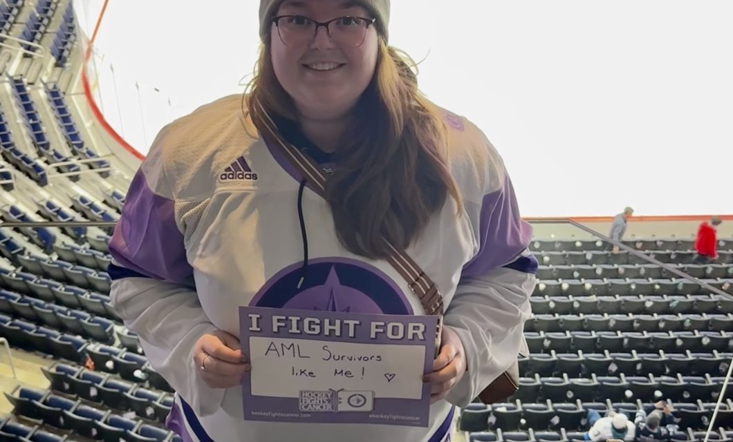 A photo of Michaela standing inside of a hockey arena, smiling, wearing a grey hat and a purple "hockey fights cancer" Winnipeg Jets jersey, holding a sign that says "I fight for AML survivors like me."