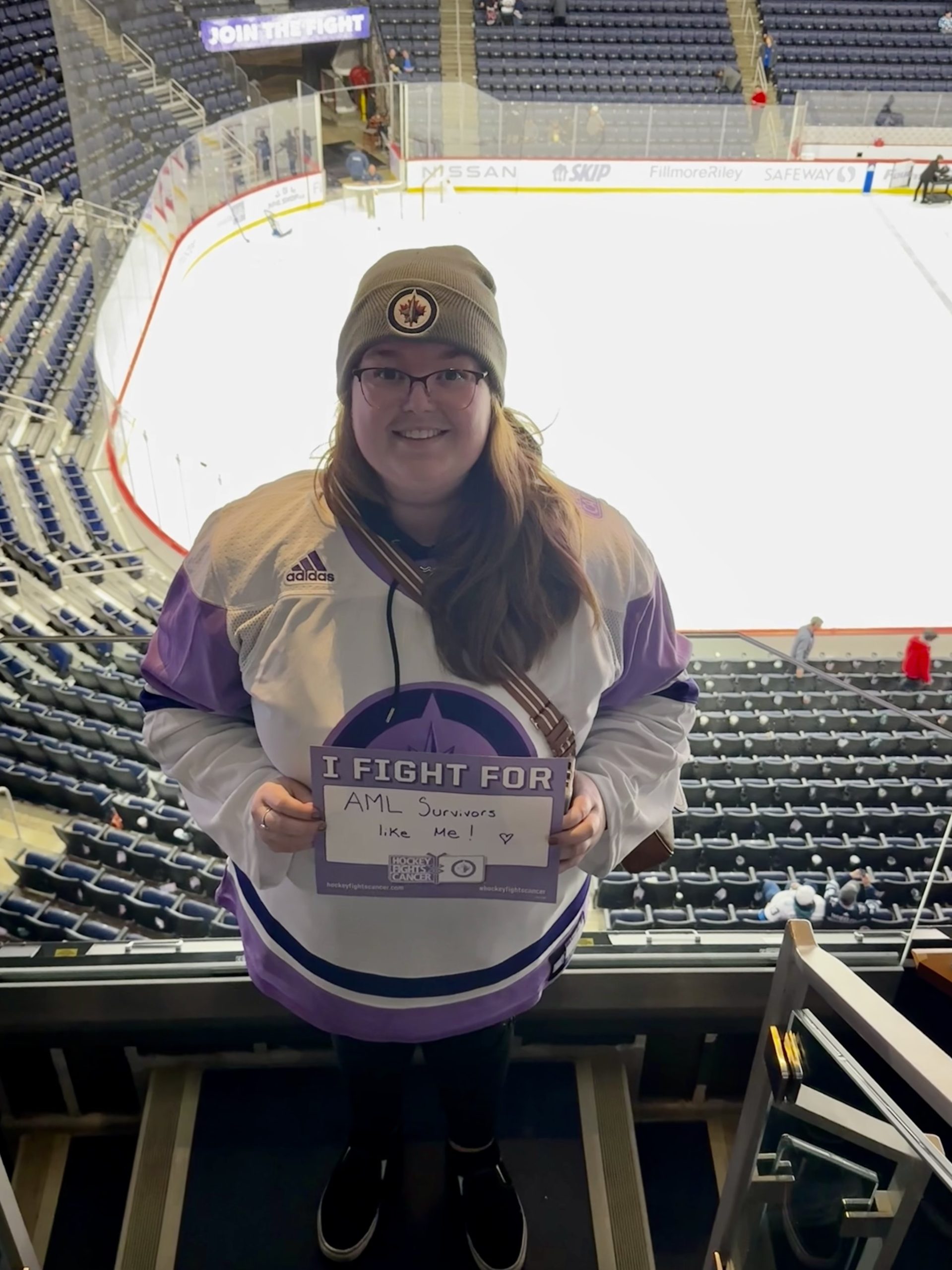 A photo of Michaela standing inside of a hockey arena, smiling, wearing a grey hat and a purple "hockey fights cancer" Winnipeg Jets jersey, holding a sign that says "I fight for AML survivors like me."