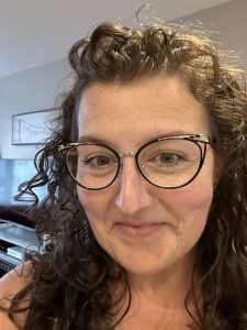 A close-up selfie of Sarah smiling at the camera. Sarah is a white women in her early 40s. Sarah has long dark brown curly hair, an oval shaped face and wears cat eye-shaped glasses.