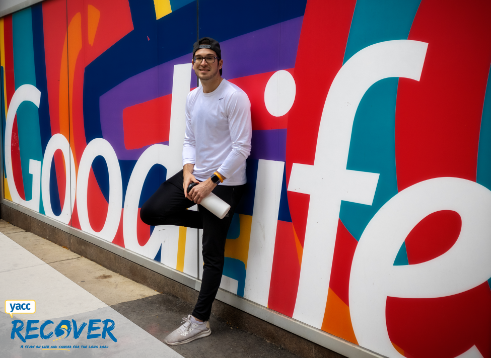 Alex leans against the wall of a GoodLife gym. There is a colourful GoodLife sign covering the wall. He is holding a water bottle. 