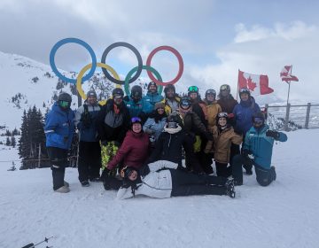 A group of 13 members of the YACC community and four snow sports instructors stand in front of the Olympic rings on Whistler mountain dressed in winter sports gear and smiling