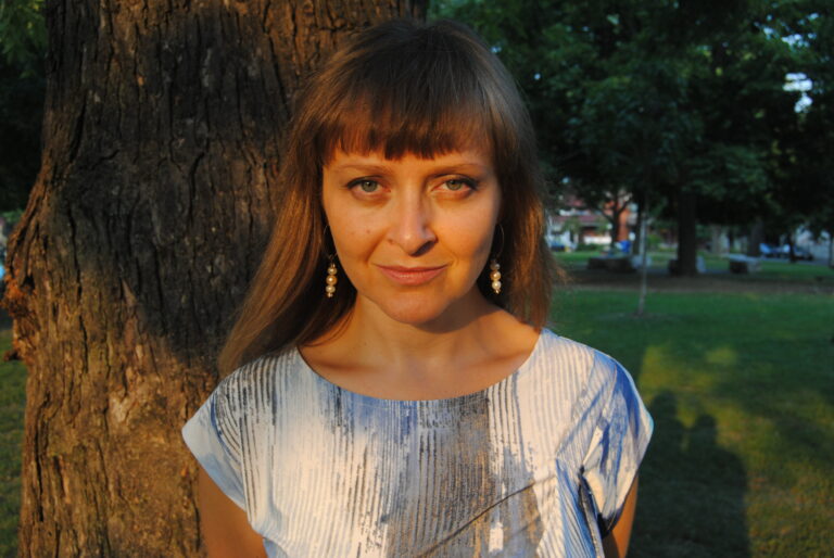 A mid-range photo of Teva staring straight at the camera. She is outside and you can see a tree behind her. She is wearing a white shirt and earrings, and she has bangs.