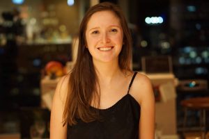 A medium-wide shot of Hannah smiling at the camera. Her posture is relaxed. She is wearing a black tank top and her long brown hair is down. 