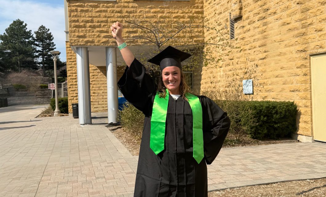 A full-body shot of Krista with her fist held over her head in victory, wearing a graduation cap and gown, outside of her cancer clinic.