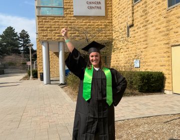 A full-body shot of Krista with her fist held over her head in victory, wearing a graduation cap and gown, outside of her cancer clinic.