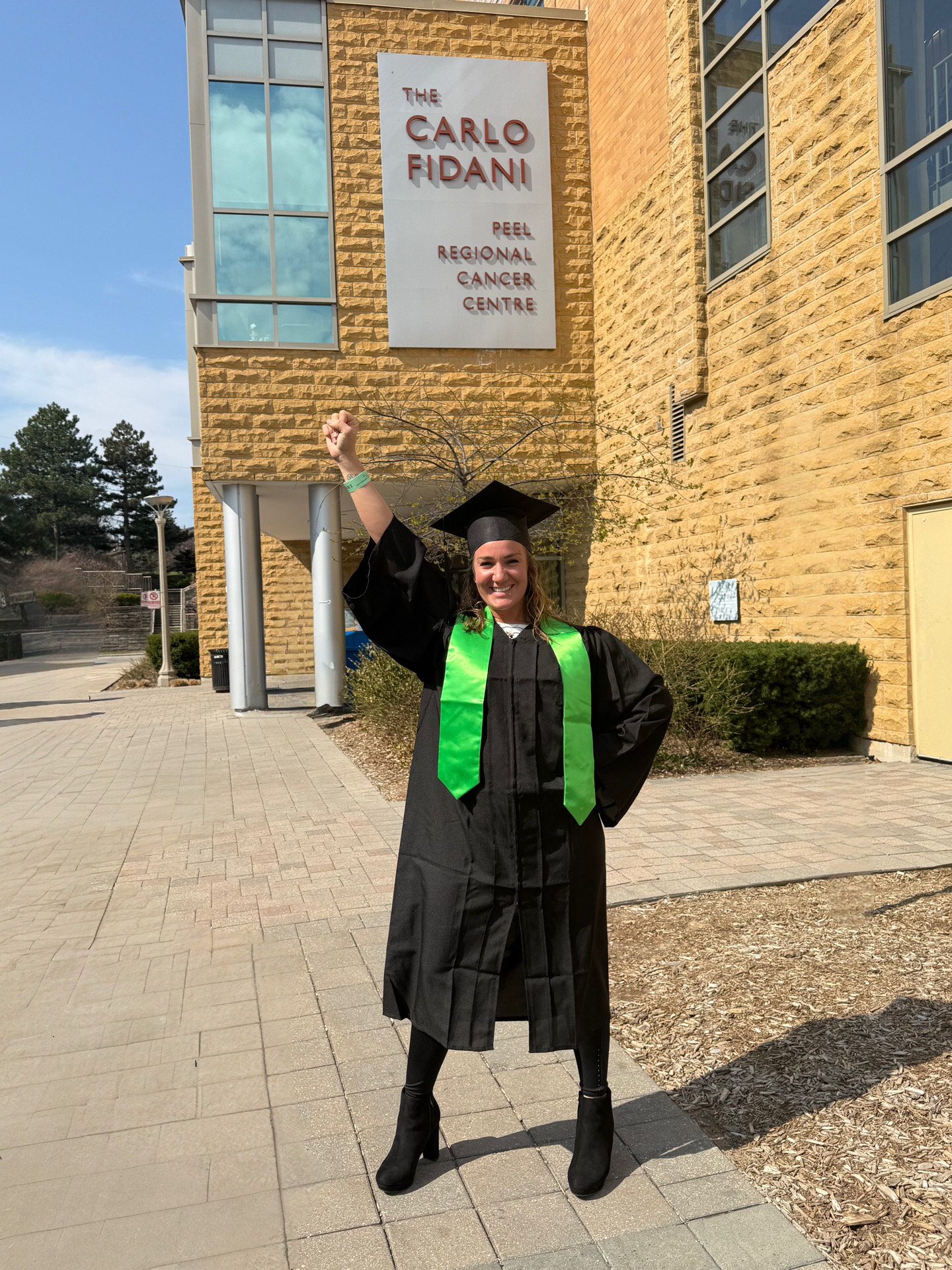 A full-body shot of Krista with her fist held over her head in victory, wearing a graduation cap and gown, outside of her cancer clinic. 
