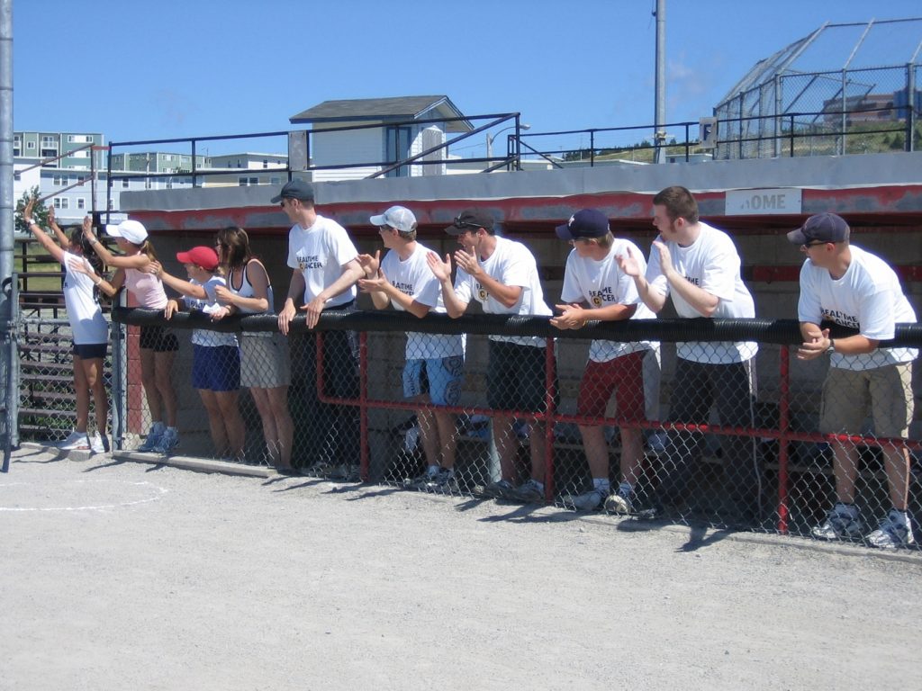 A softball team lines a fence by the dugout, cheering for their teammate. 