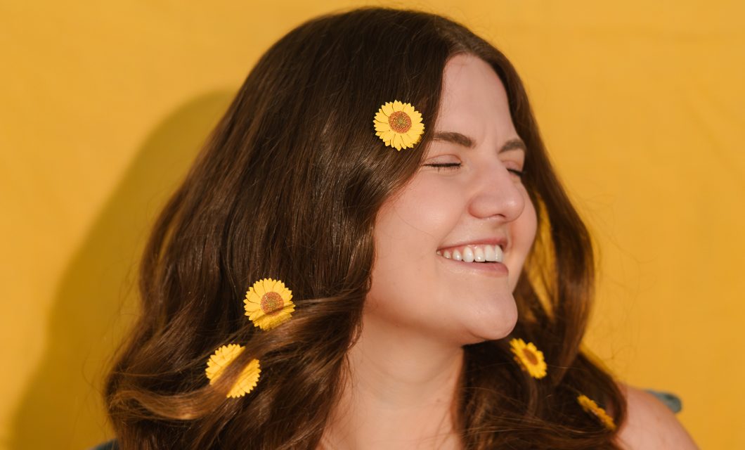 A medium headshot of Emily with small sunflowers in her long brown hair. She is wearing a sleeveless green shirt.