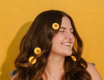 A medium headshot of Emily with small sunflowers in her long brown hair. She is wearing a sleeveless green shirt.