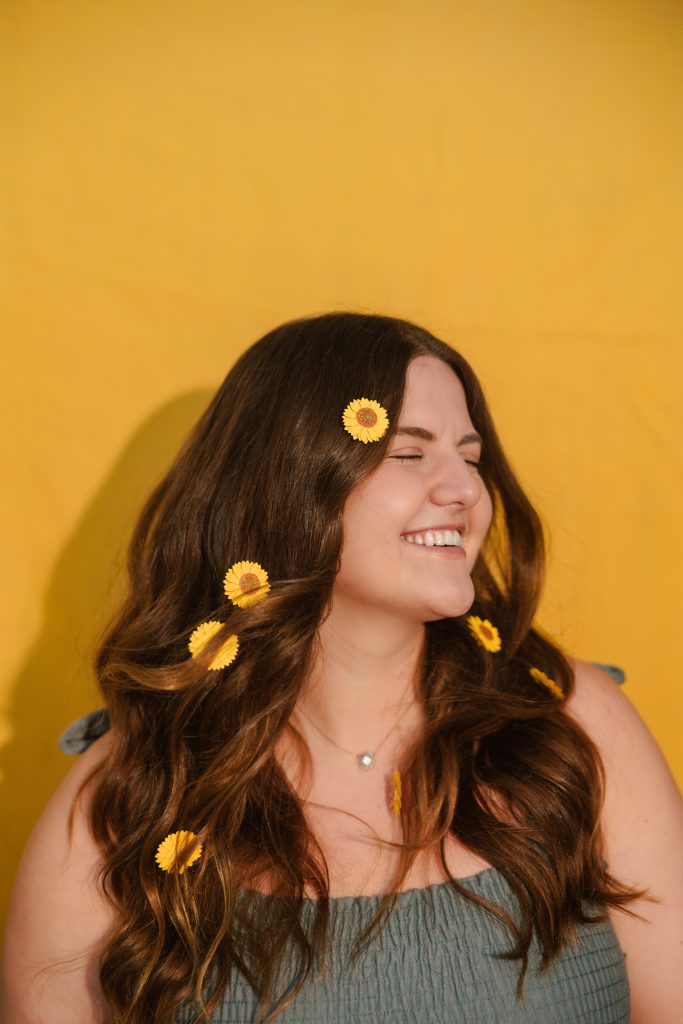 A medium headshot of Emily with small sunflowers in her long brown hair. She is wearing a sleeveless green shirt.