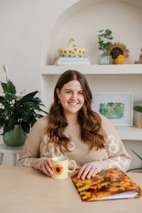 A professional headshot of Emily sitting at a desk, holding a white mug with a sunflower on it. 