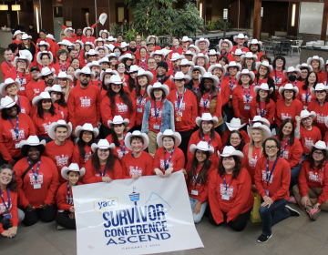 A group photo of over 100 young adults smiling at the camera in the hotel lobby. They are all wearing red shirts with a white conference logo on them, and most are wearing white cowboy hats from the Calgary Stampede.