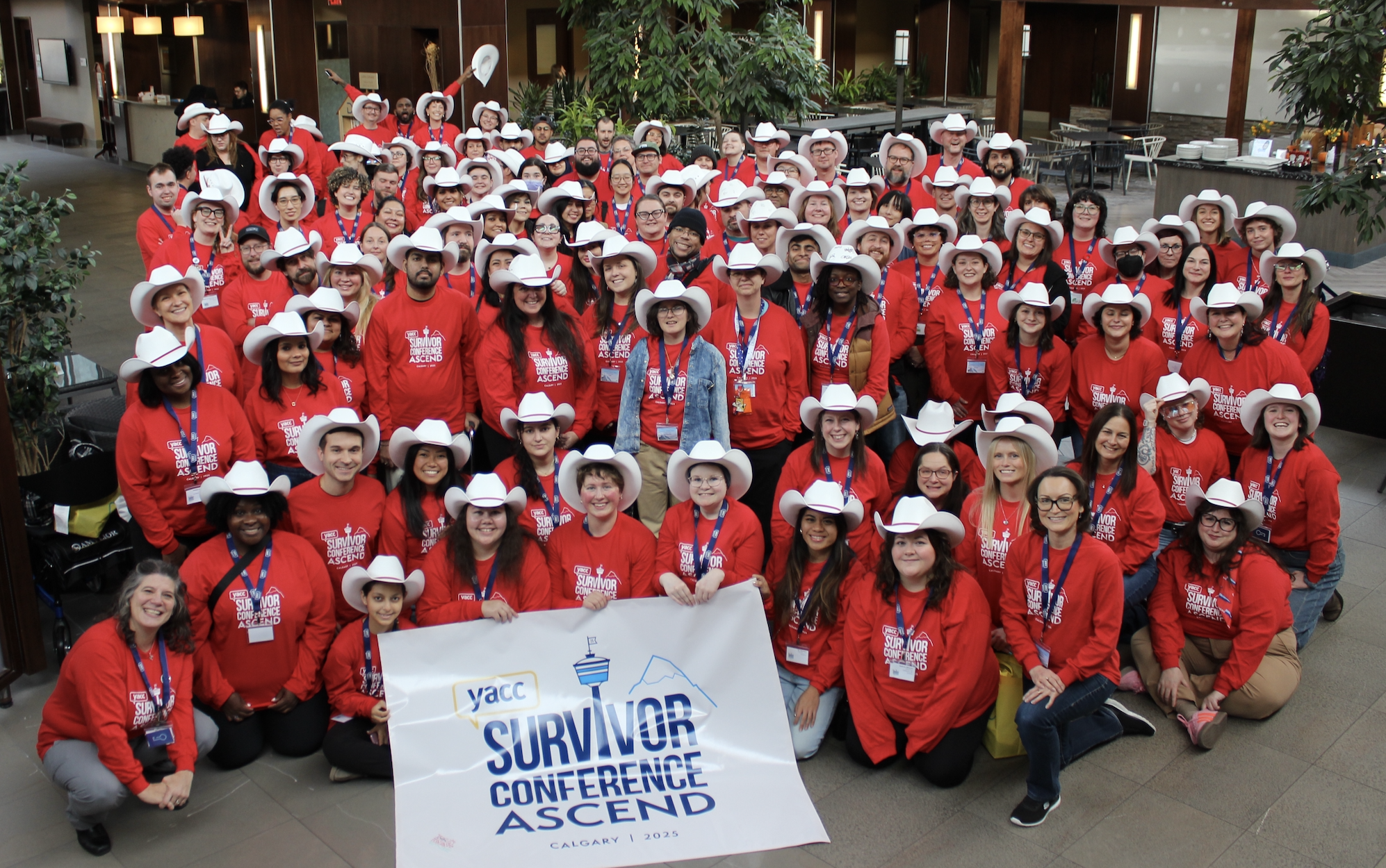 A group photo of over 100 young adults smiling at the camera in the hotel lobby. They are all wearing red shirts with a white conference logo on them, and most are wearing white cowboy hats from the Calgary Stampede.