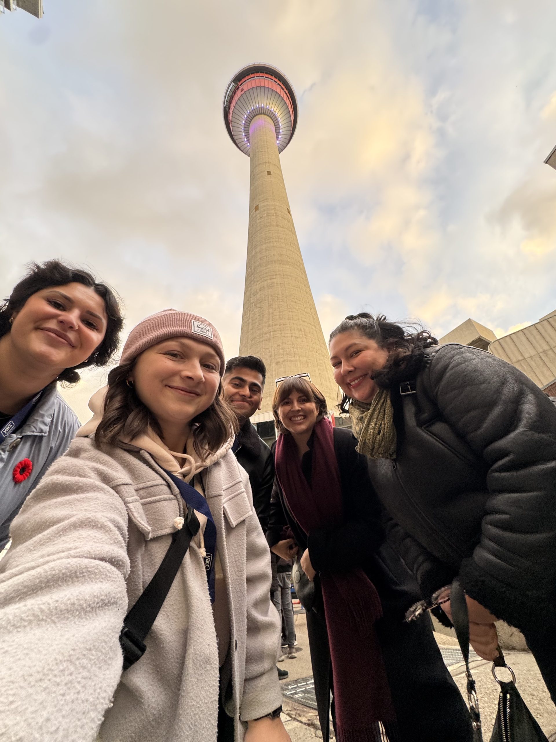 Five young adults smile down at the camera in a group selfie featuring the Calgary Tower above them.