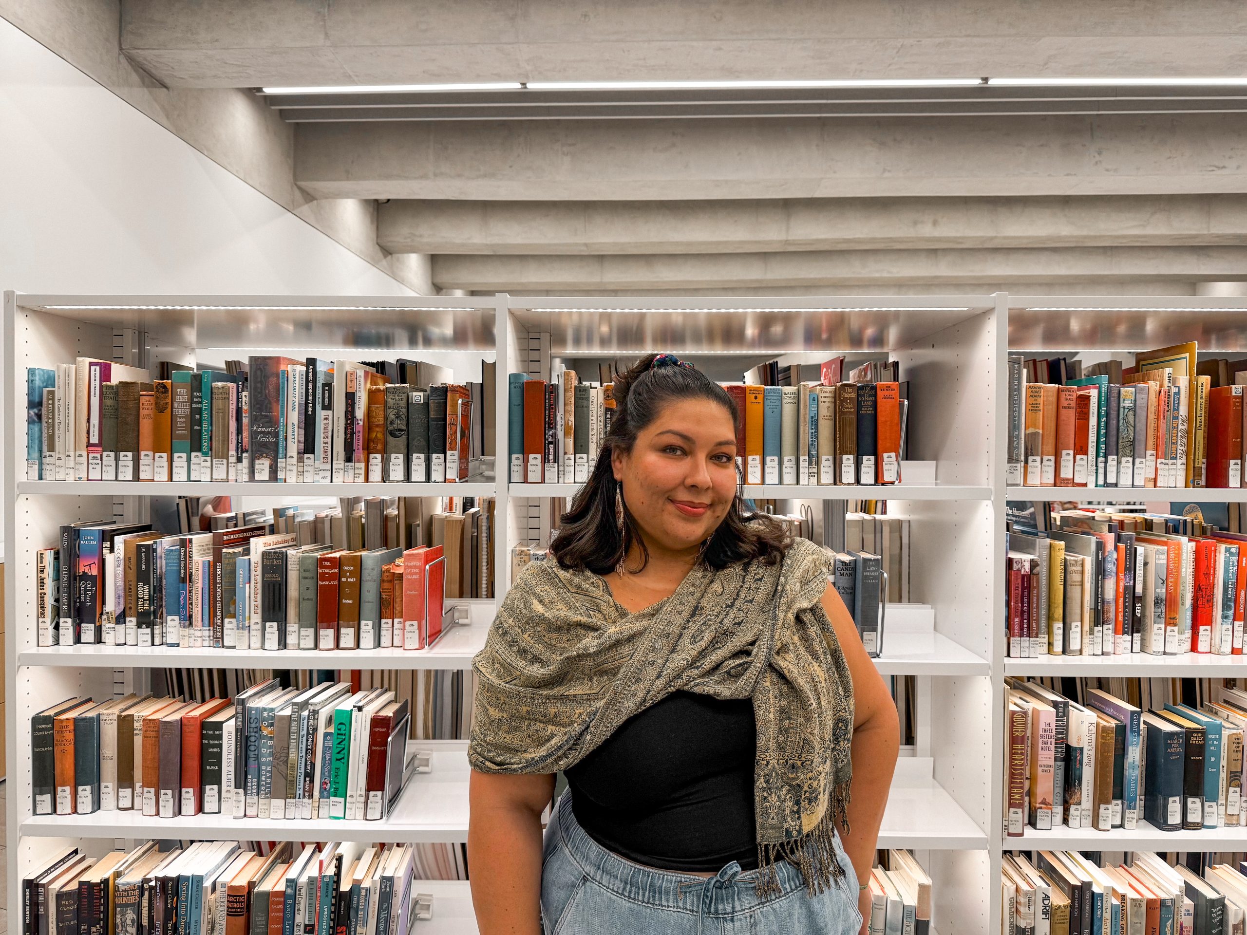 In this medium-wide shot, Dani poses in front of a book stack in a library. She is wearing light blue jeans, a black shirt, a neutral-coloured scarf around her shoulders, and is wearing her shoulder-length hair in a half up, half down style.
