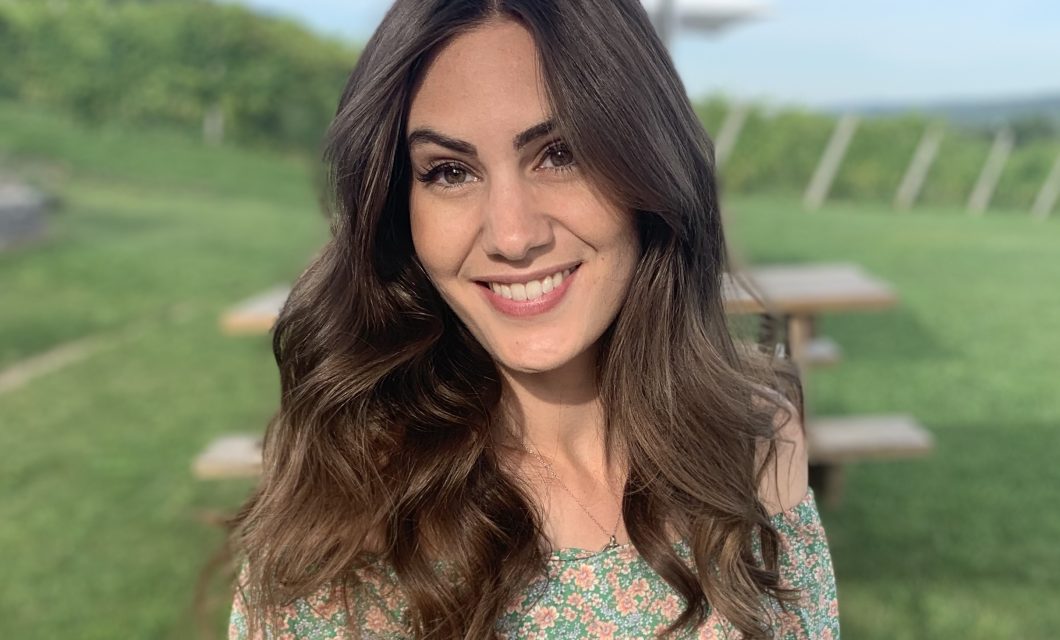 A medium shot of Ashley outside with a grassy background. She is wearing a light green dress, and her medium-length brown hair is in loose waves.