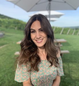 A medium shot of Ashley outside with a grassy background. She is wearing a light green dress, and her medium-length brown hair is in loose waves. 
